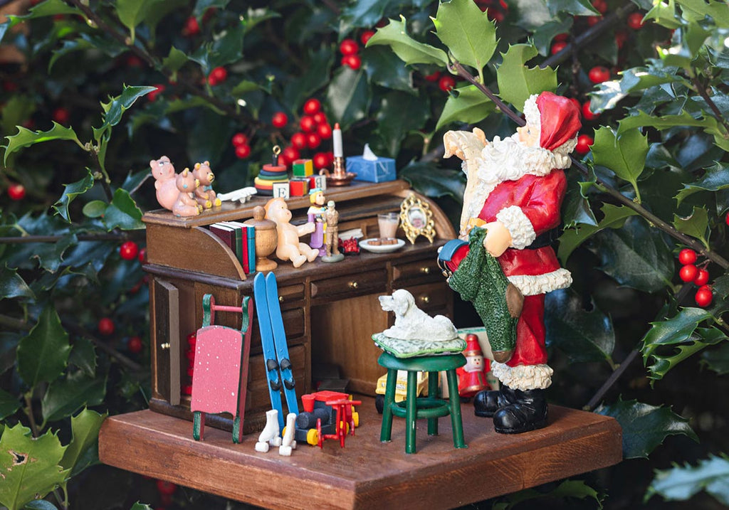 Miniature Santa Claus workshop diorama in front of a green bush. Toys, candles and cookies are on the desk and Santa Claus is reading the gift list.