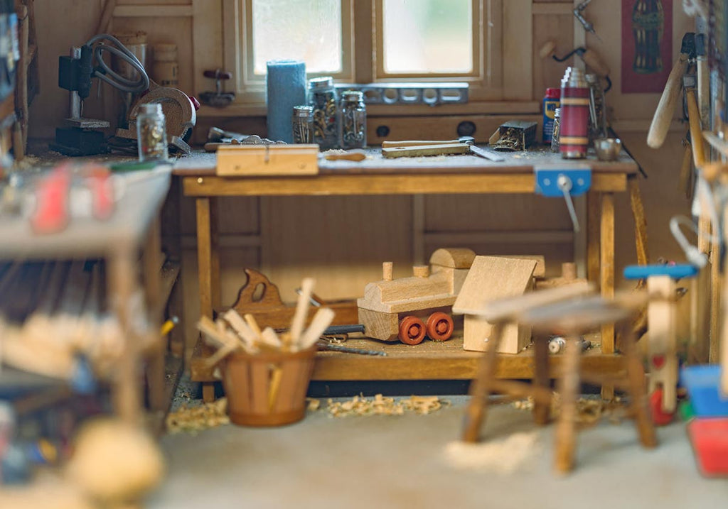 Miniature workshop garage with woodscraps and tools on the table. A miniature wooden toy train is under the table, behind the wooden stool.